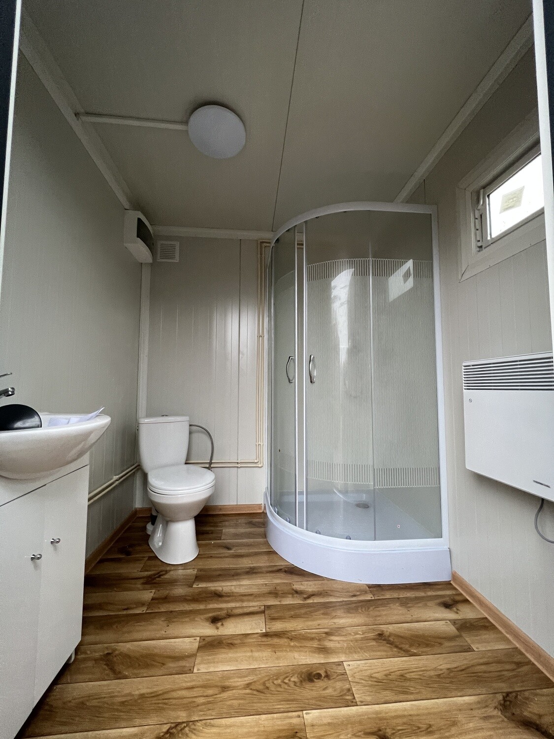 Innenansicht mit Waschbecken, WC und Rundduschkabine auf Holzdekor-Boden, Wandheizung und Fenster