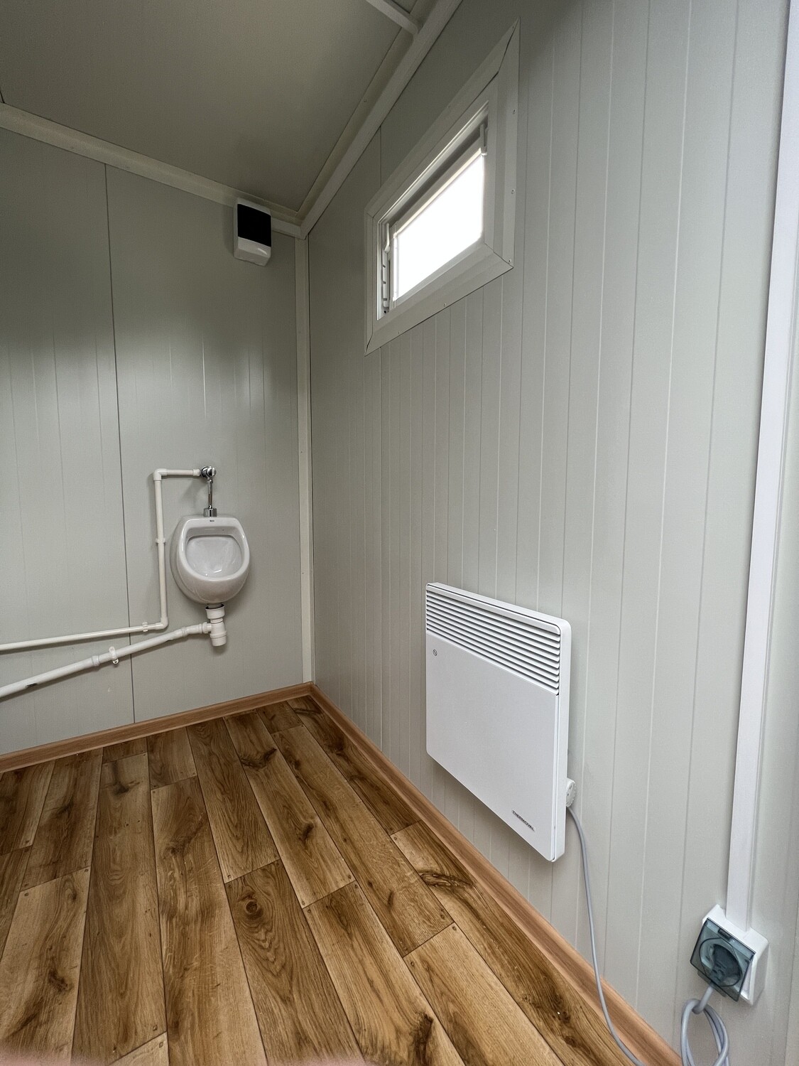 Innenraum des WC Containers: Urinal mit Aufputzleitung, Fenster, Wandkonvektor und Steckdose