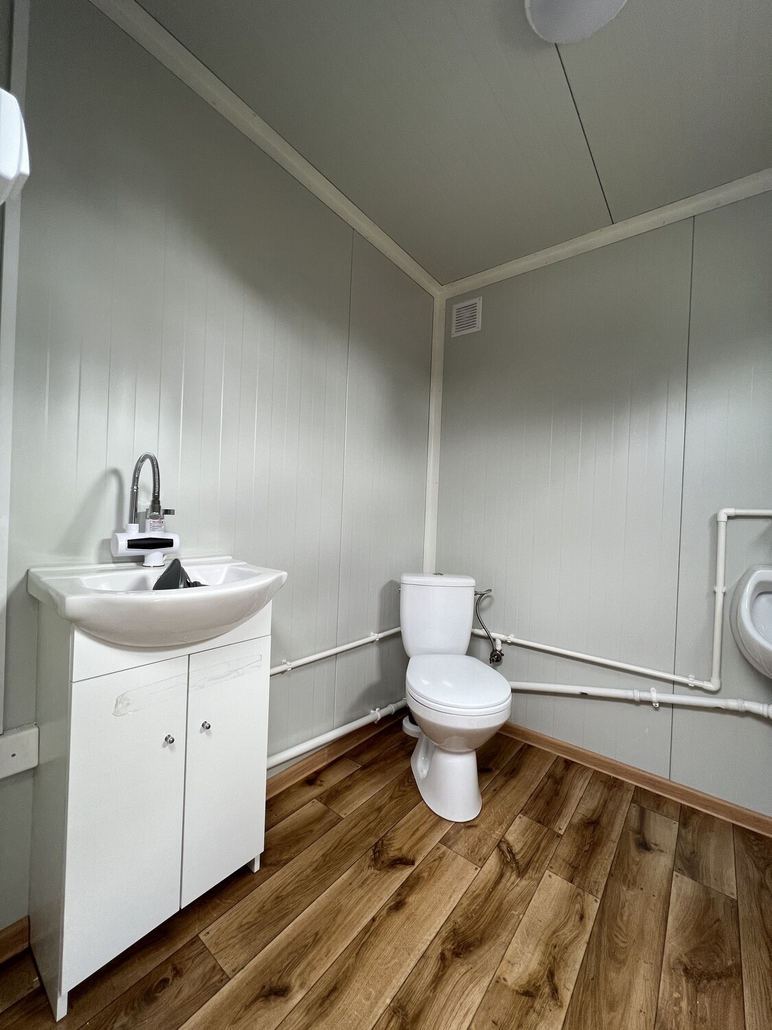 Blick auf Waschtischunterschrank, WC und Urinal im Sanitärraum mit Holzdekor-Boden und Lüftungsgitter