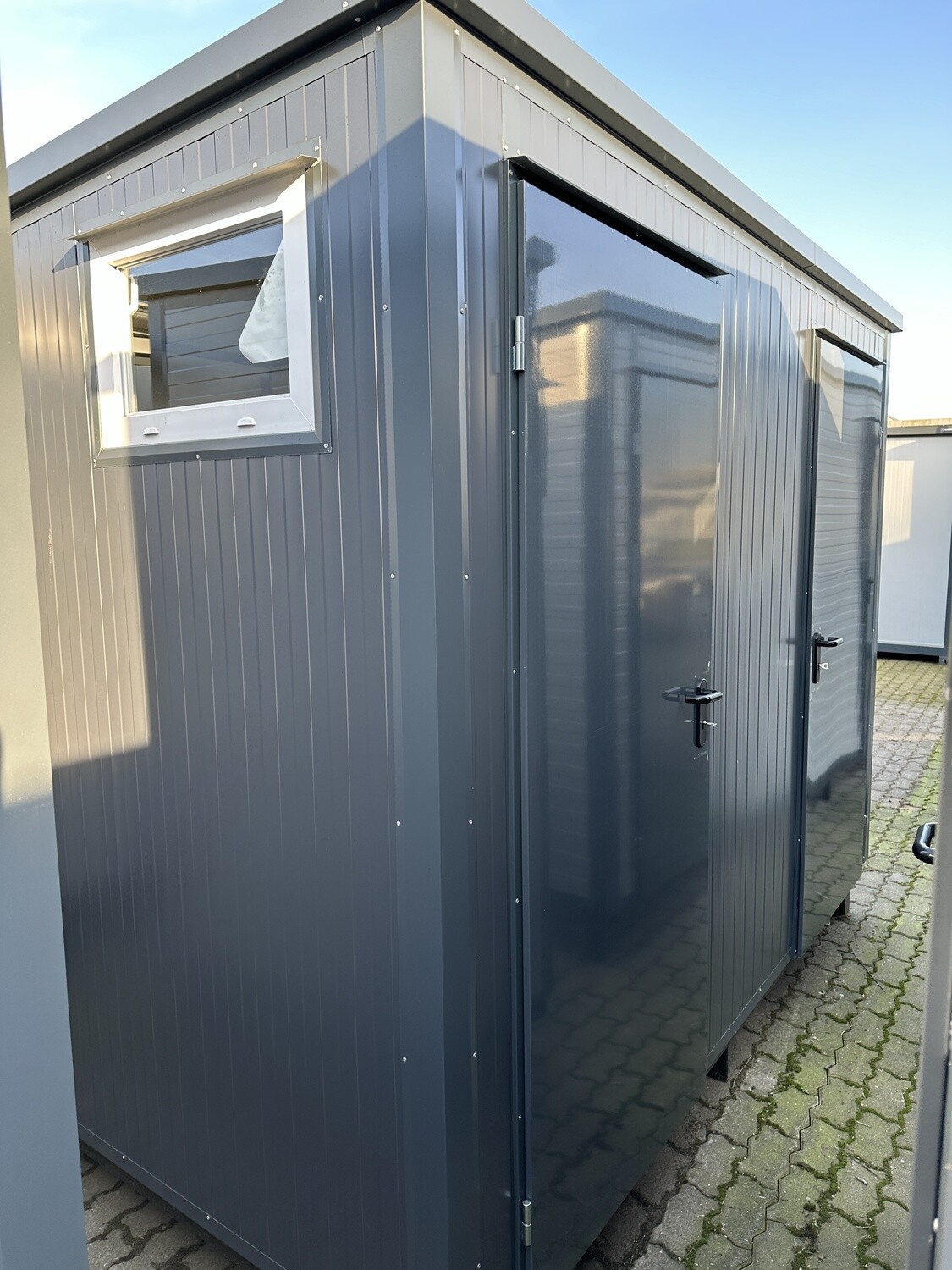 Schrägansicht auf die geöffnete Tür und das gekippte Fenster des Doppel-WC Containers auf gepflastertem Platz