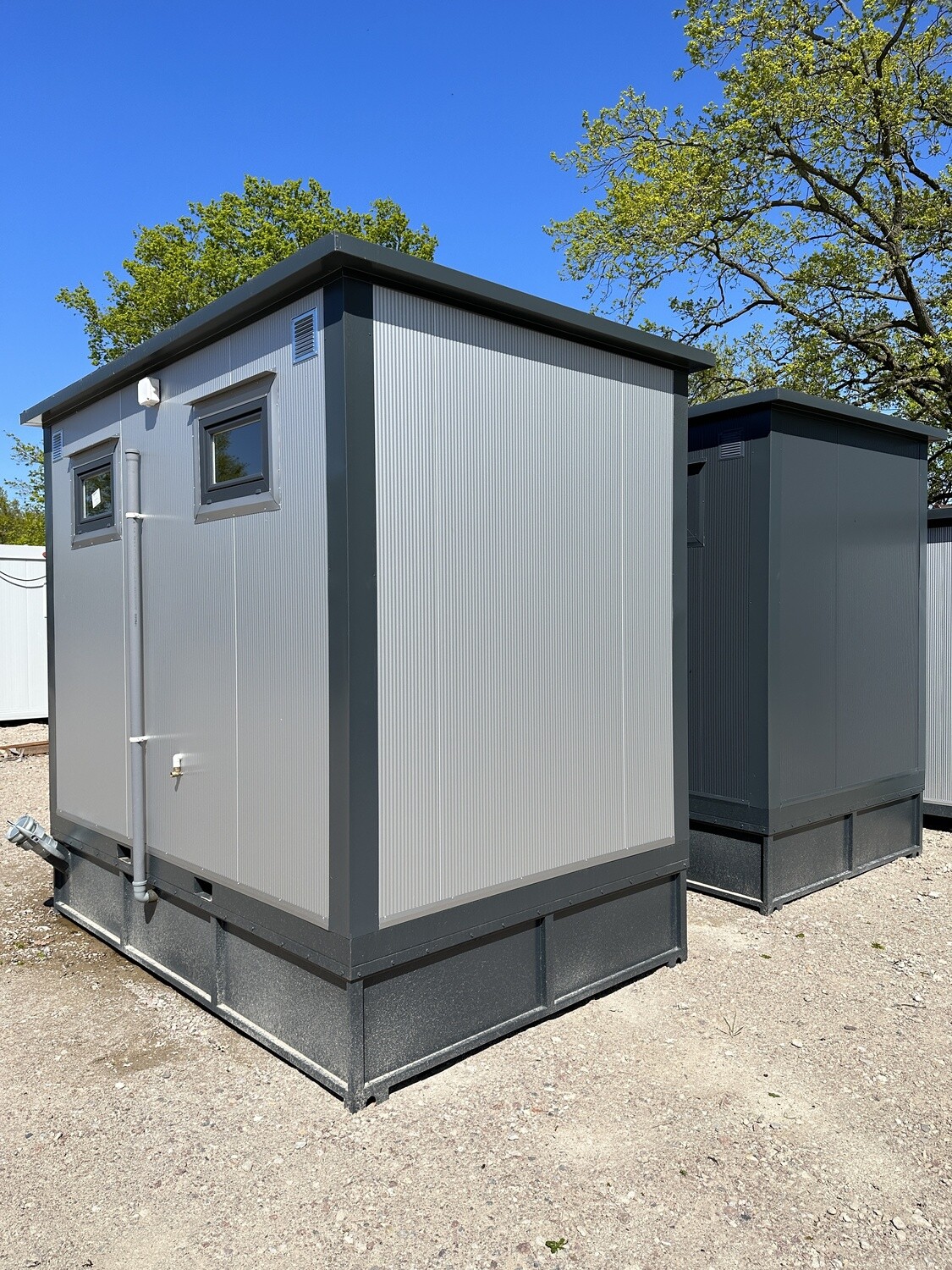 Seitenansicht eines grauen WC Containers mit kleinen Fenstern und Fallrohr, dahinter ein zweiter Container