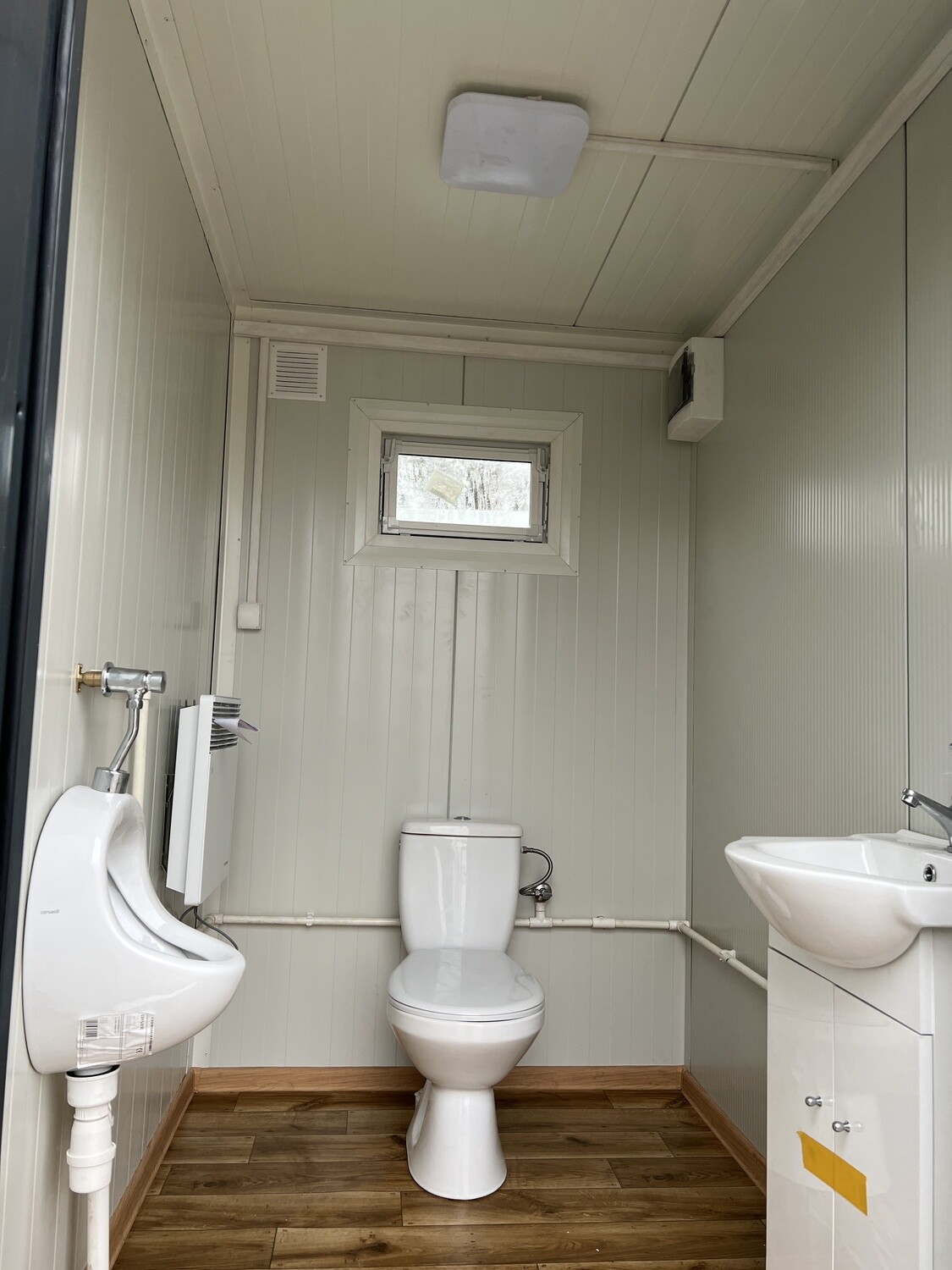 Kabine mit Urinal und WC über holzoptischem Bodenbelag, rechts Waschtisch mit weißem Unterschrank