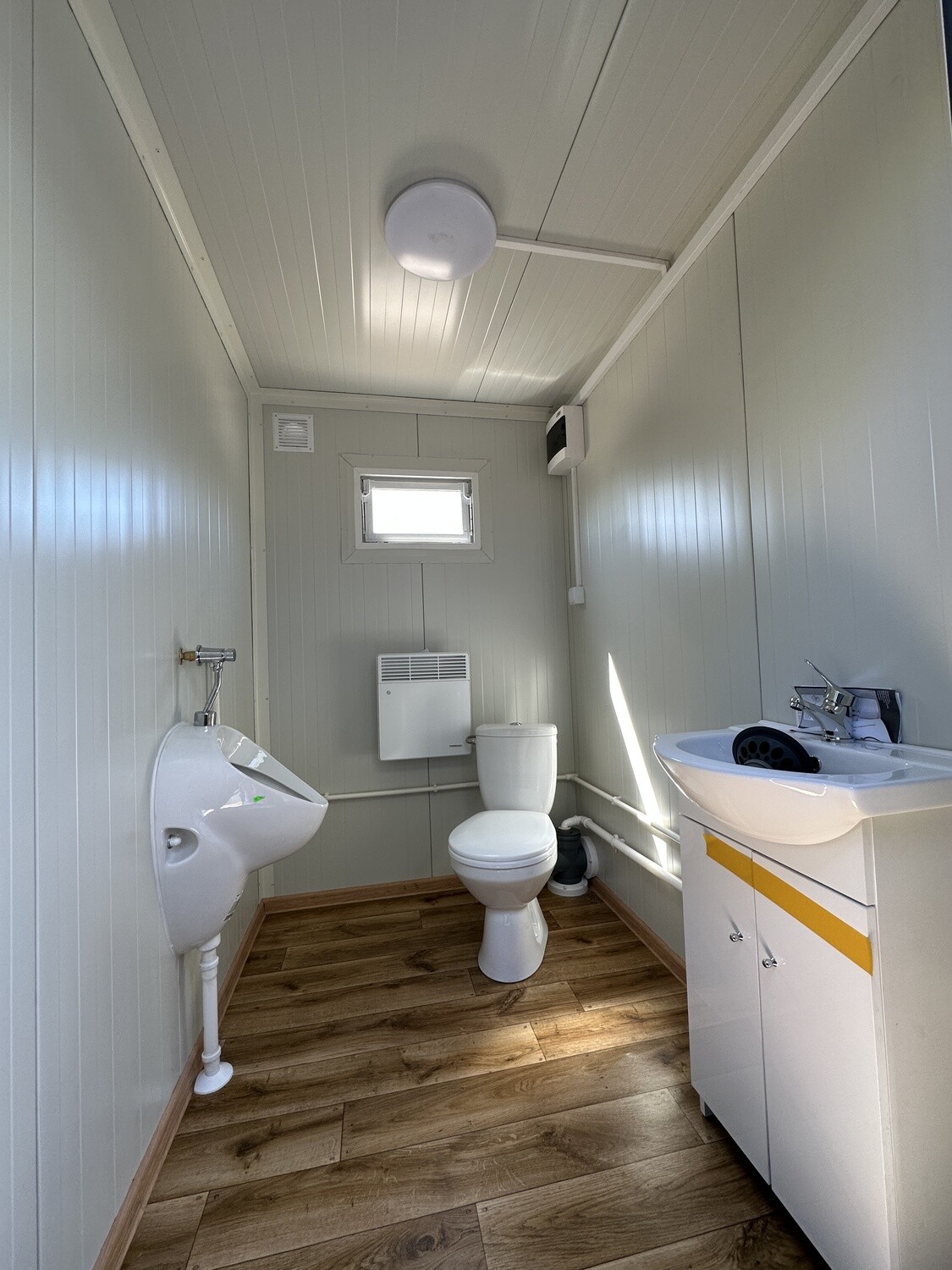 Innenraum mit Urinal, Toilette, Heizung unter dem Fenster und Waschtisch, darunter Bodenbelag in Holzoptik