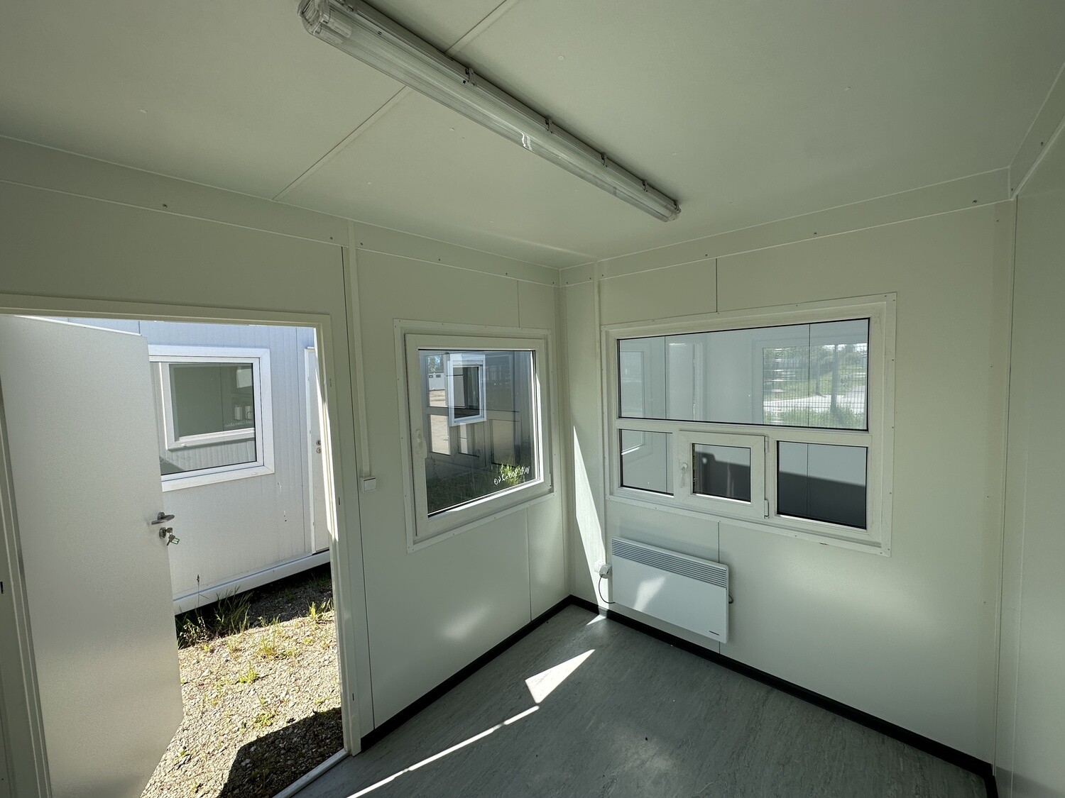 Innenraum mit Deckenleuchte, Wandheizgerät unter dem Fenster und grauem Bodenbelag im Bürocontainer