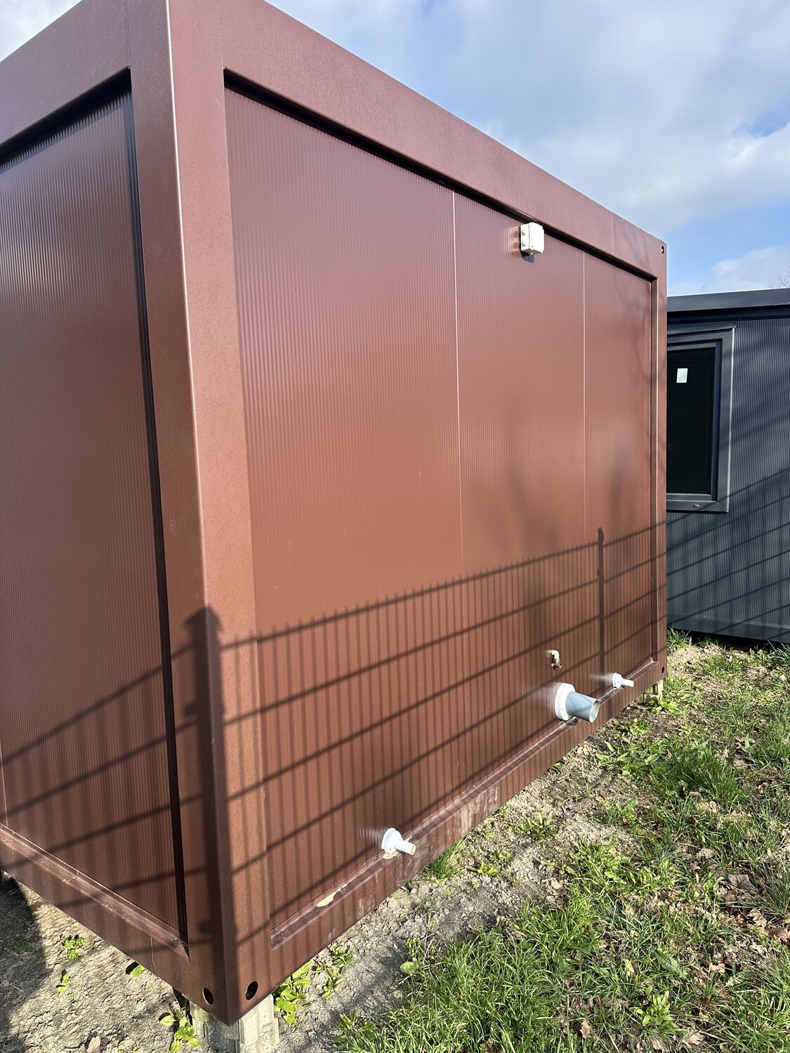 Braune Rückwand mit weißen Rohranschlüssen und Elektrokasten am Doppel-WC und Duschcontainer