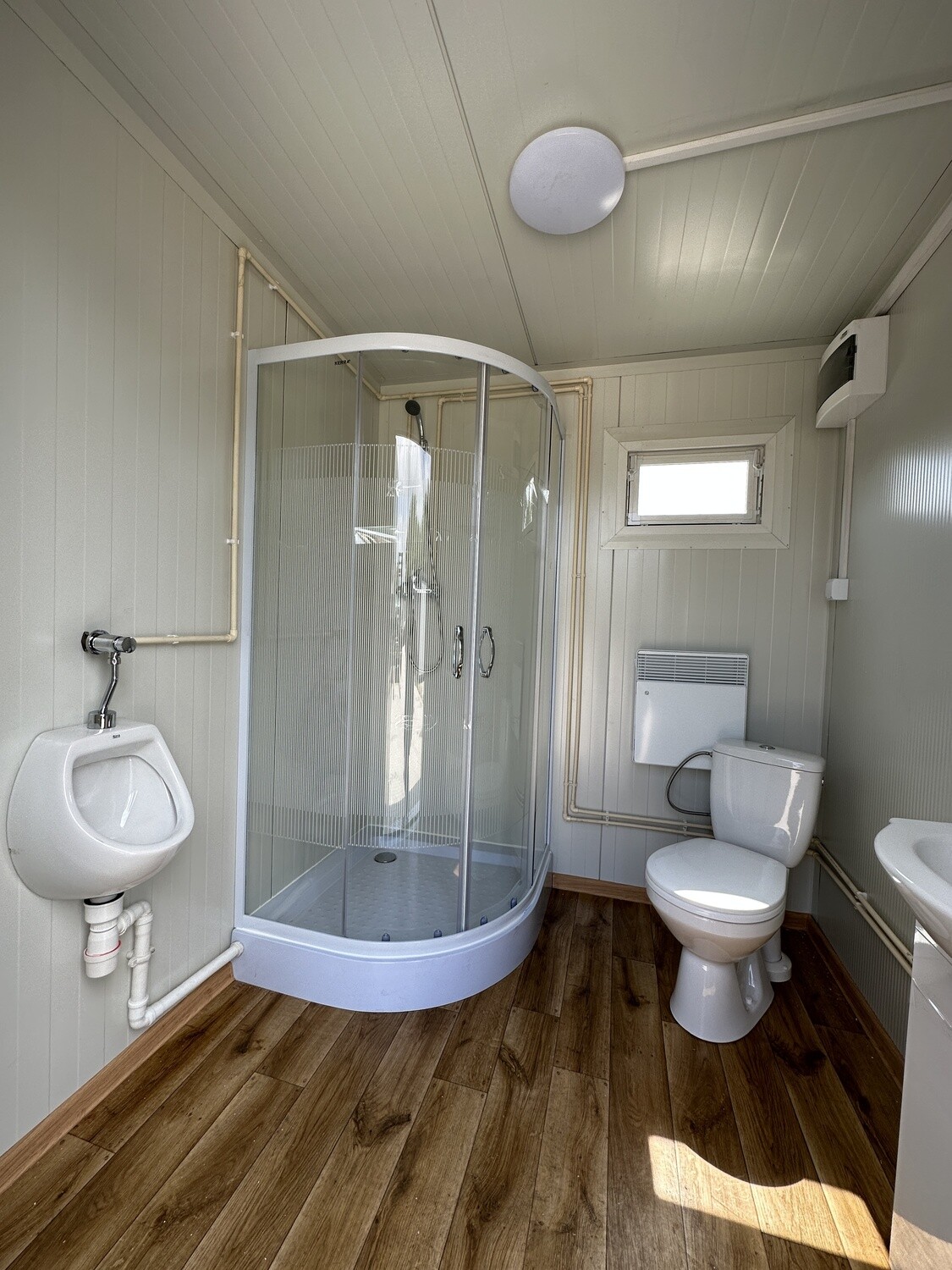 Innenraum mit Urinal, runder Duschkabine, WC und Wandheizgerät auf Boden in Holzoptik