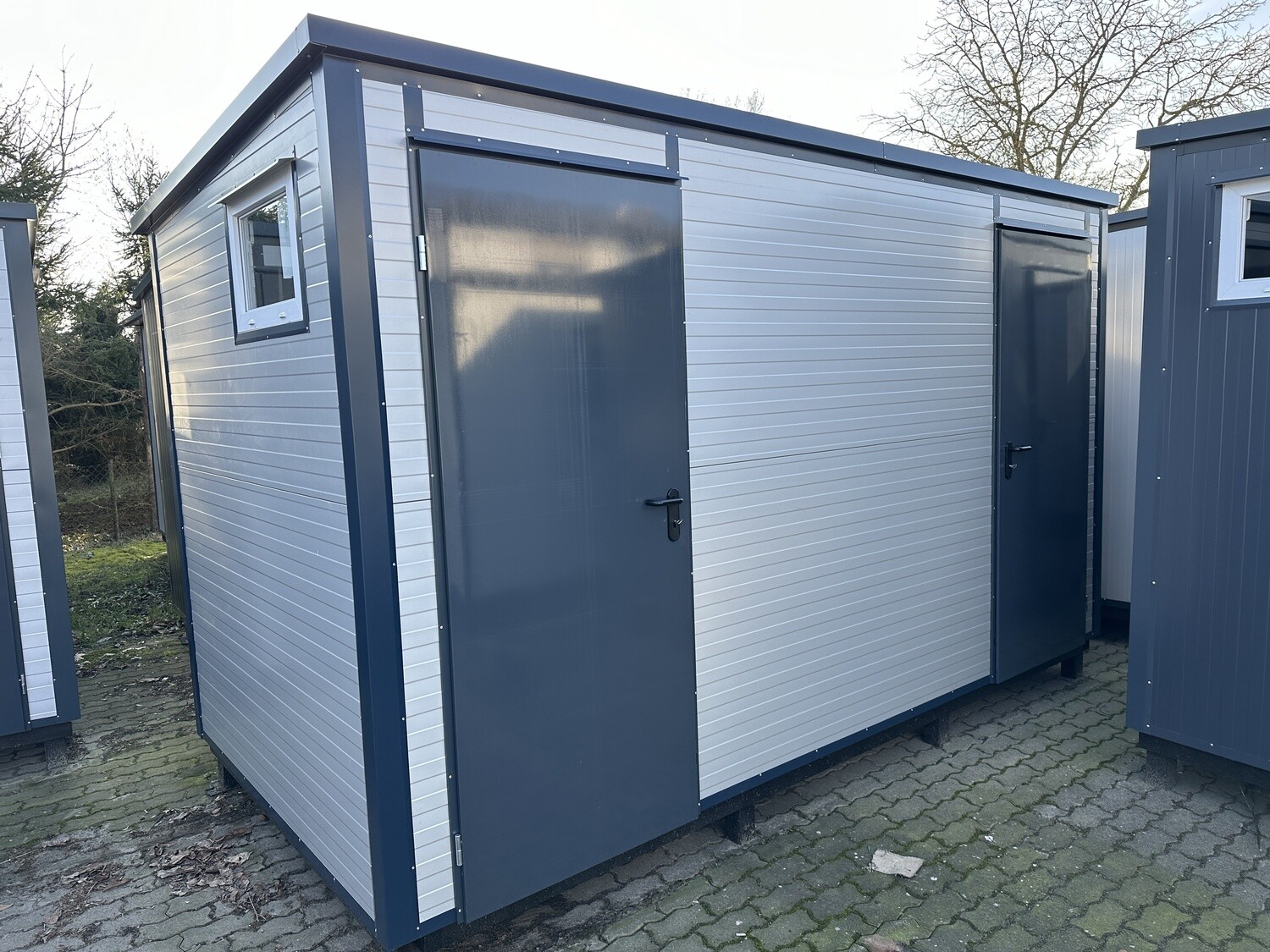 Über Eck fotografierter Sanitärcontainer mit Warmwasserboiler: helle Wandpaneele, zwei dunkle Eingangstüren