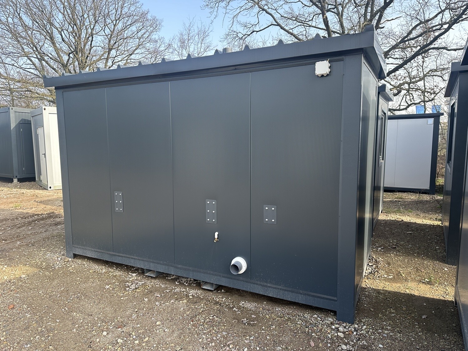 Geschlossene Längsseite des 4,00 x 2,00 m WC-Containers mit Abflussrohr und Wandkasten auf Schotter