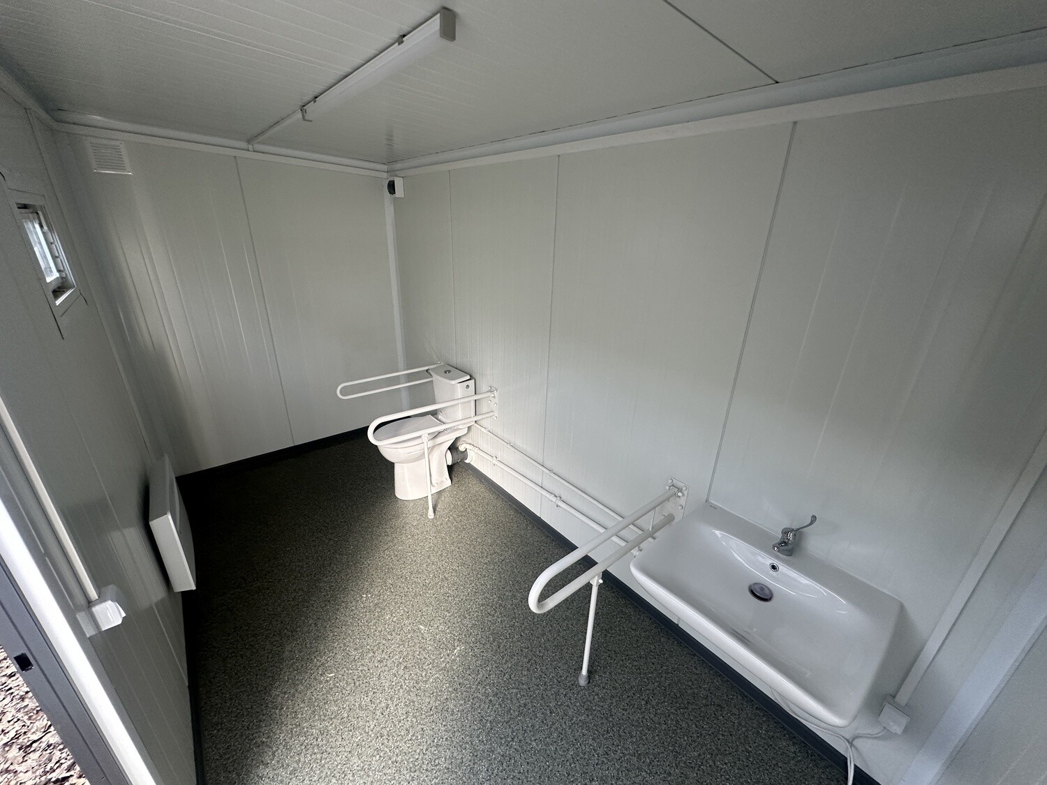 Innenansicht mit Toilette und Waschbecken samt Stützklappgriffen sowie dunklem Bodenbelag im WC-Container