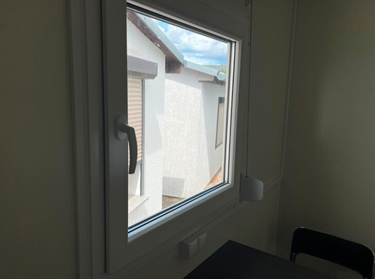 Fenster mit Griff von innen aufgenommen, daneben eine Steckdose sowie Tisch- und Stuhlkante im Büroraum
