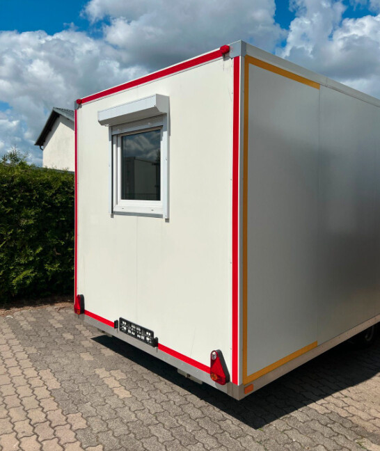 Ecke eines Büro-Anhängers mit roten Kantenprofilen, Fenster mit Rollladen und Rad auf gepflastertem Platz