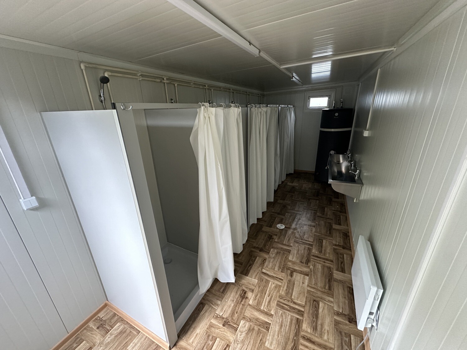 Innenansicht mit Duschkabinen hinter weißen Vorhängen, Boden in Holzoptik und Waschrinne am Ende