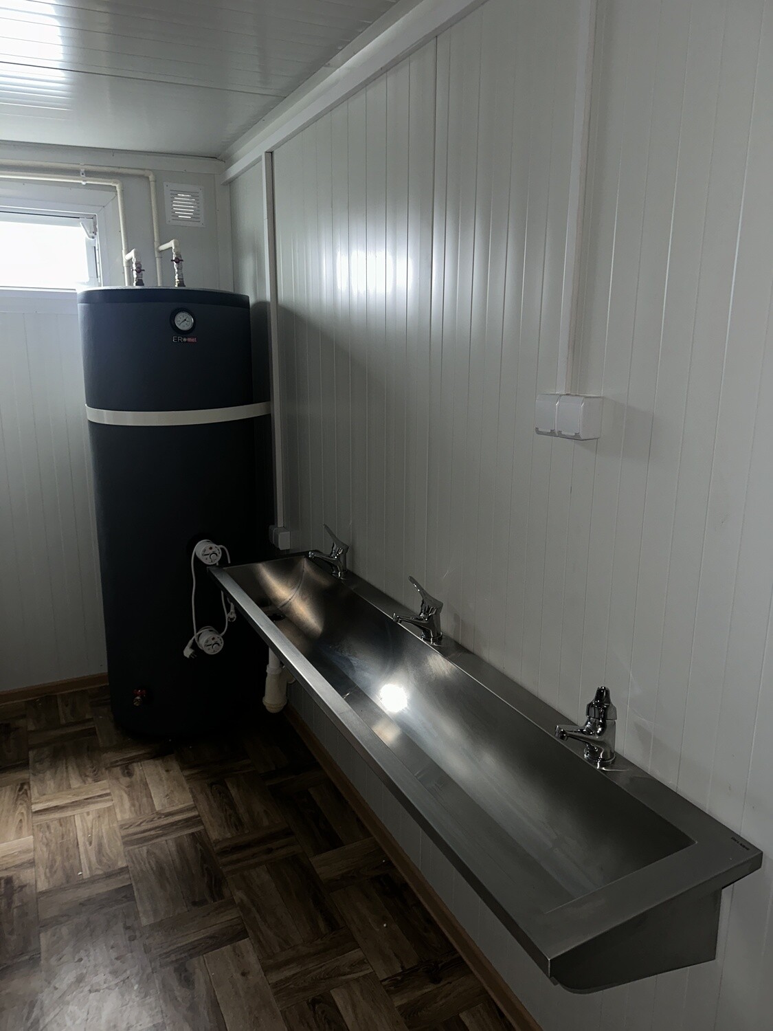 Detailaufnahme des schwarzen Warmwasserspeichers neben der Edelstahl-Waschrinne mit drei Armaturen