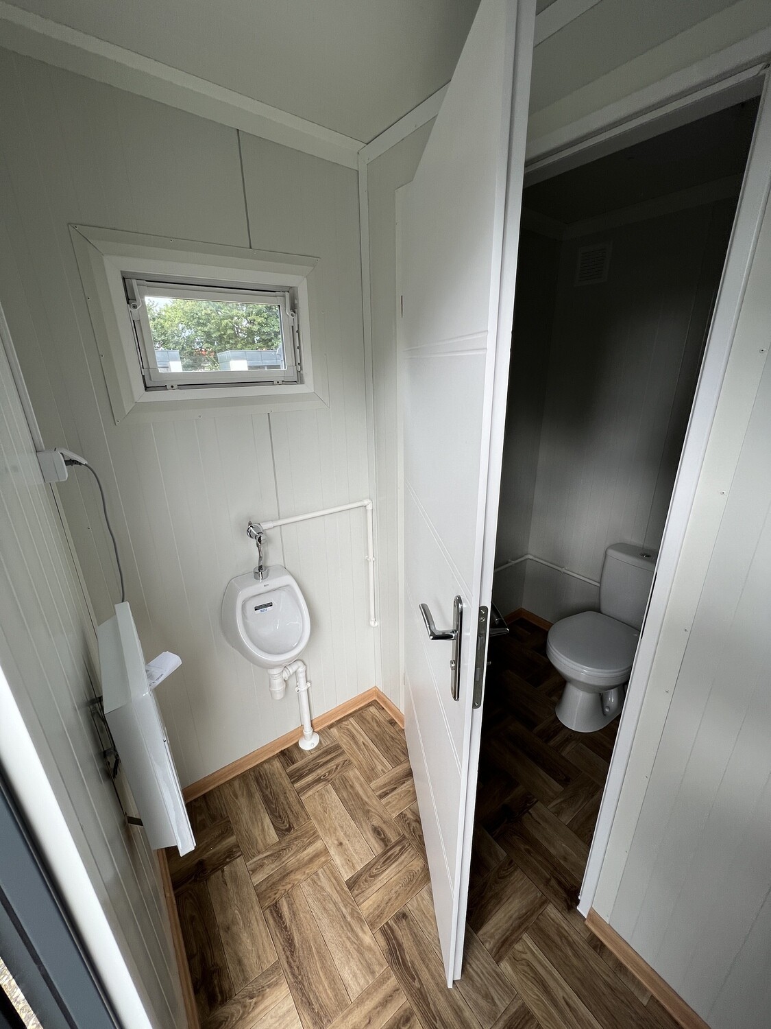 Innenraum mit wandhängendem Urinal, Kippfenster und offener Tür zur Toilettenkabine dahinter