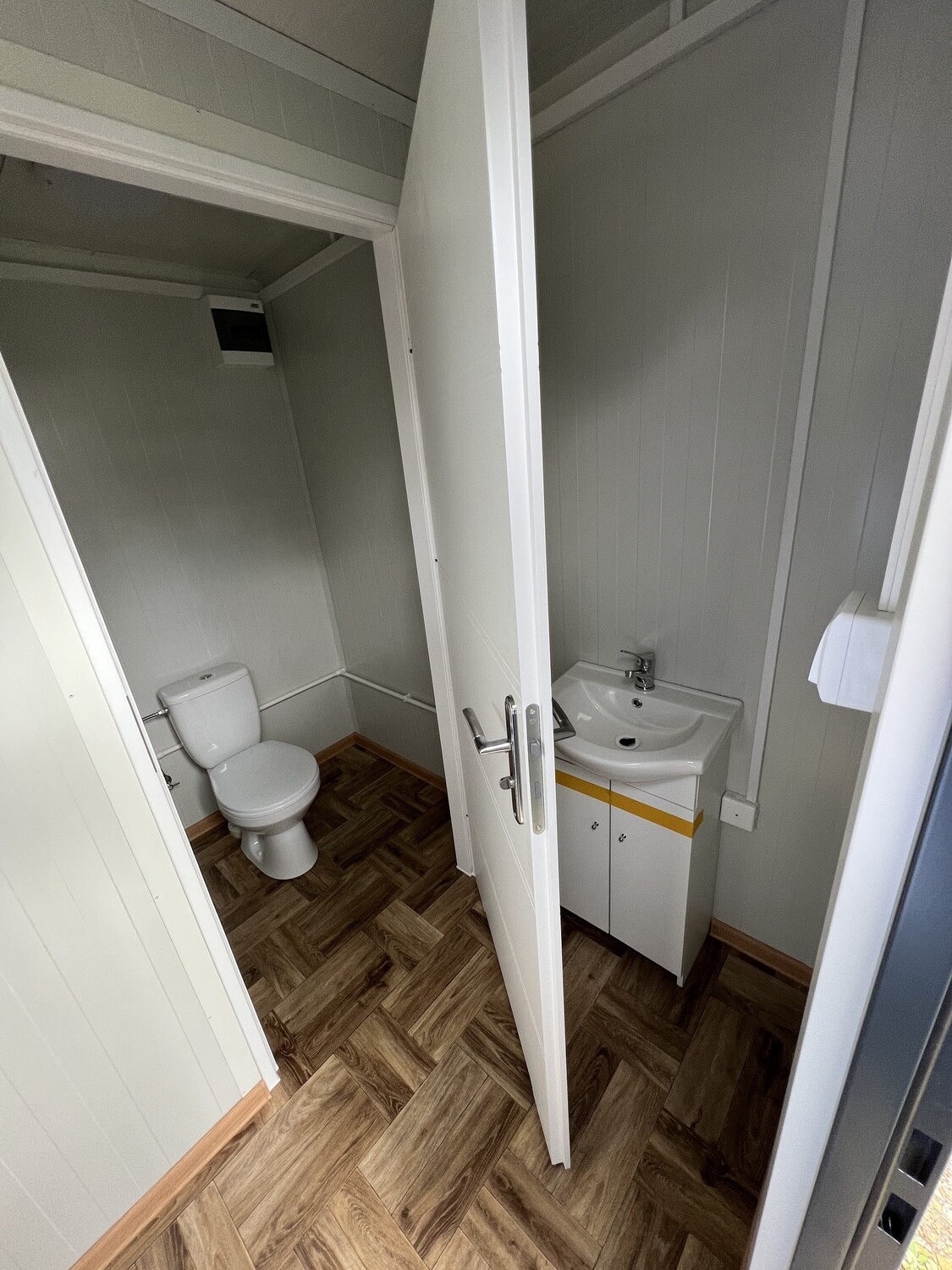 Blick auf Waschtisch mit Unterschrank und die benachbarte WC-Kabine über dem Boden in Holzoptik