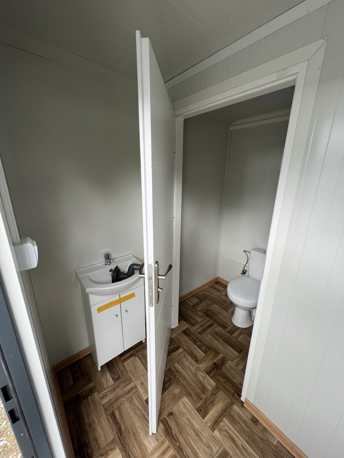 Geöffnete Kabinentür gibt den Blick auf das WC frei, links Waschbecken mit weißem Unterschrank