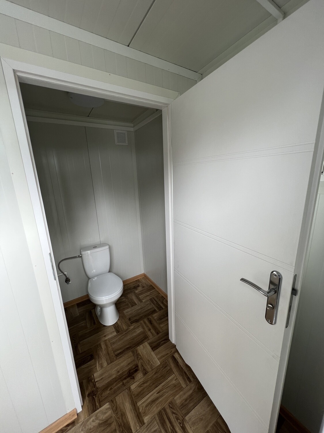 Weiße Tür mit Edelstahlklinke vor der Toilettenkabine mit Deckenleuchte und Lüftungsgitter