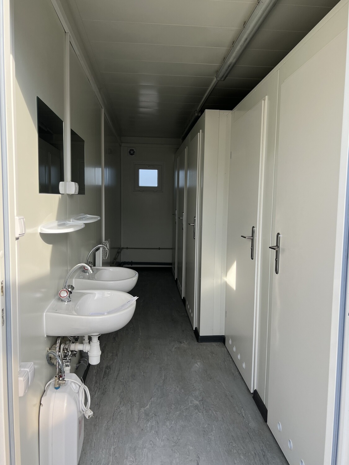 Blick durch den Mittelgang: links Waschbecken, rechts die Türen der Toilettenkabinen, Fenster hinten