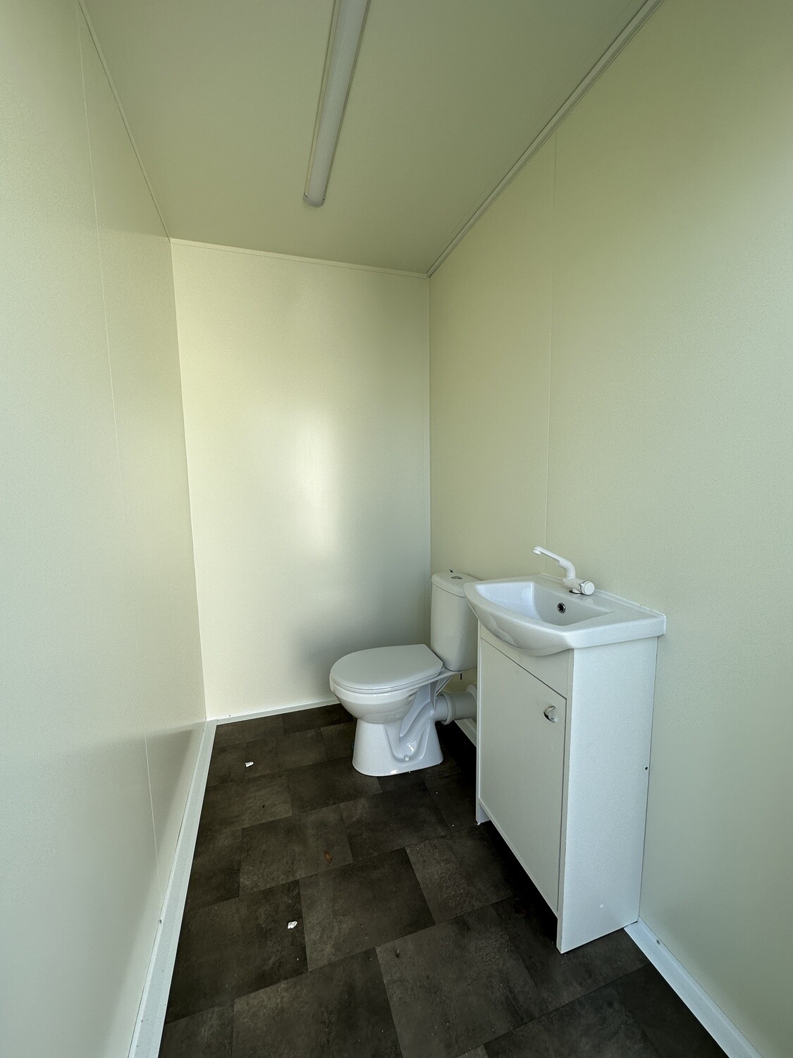 Innenraum mit Stand-WC, Waschtisch mit Unterschrank und Deckenleuchte über dunklem Bodenbelag