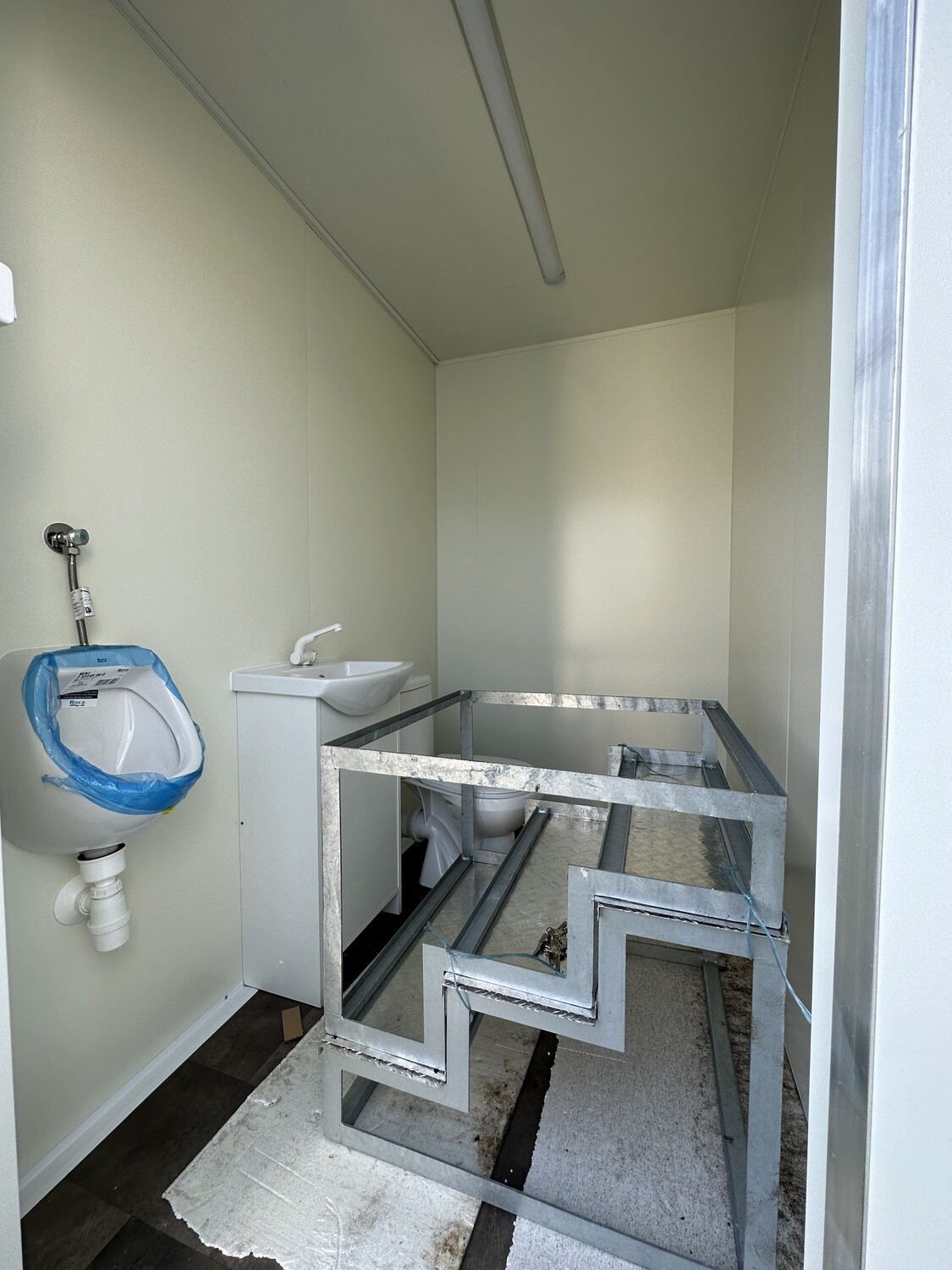 Noch im Aufbau: Urinal mit Schutzfolie, Stahlrahmen auf dem unfertigen Boden neben dem Waschtisch