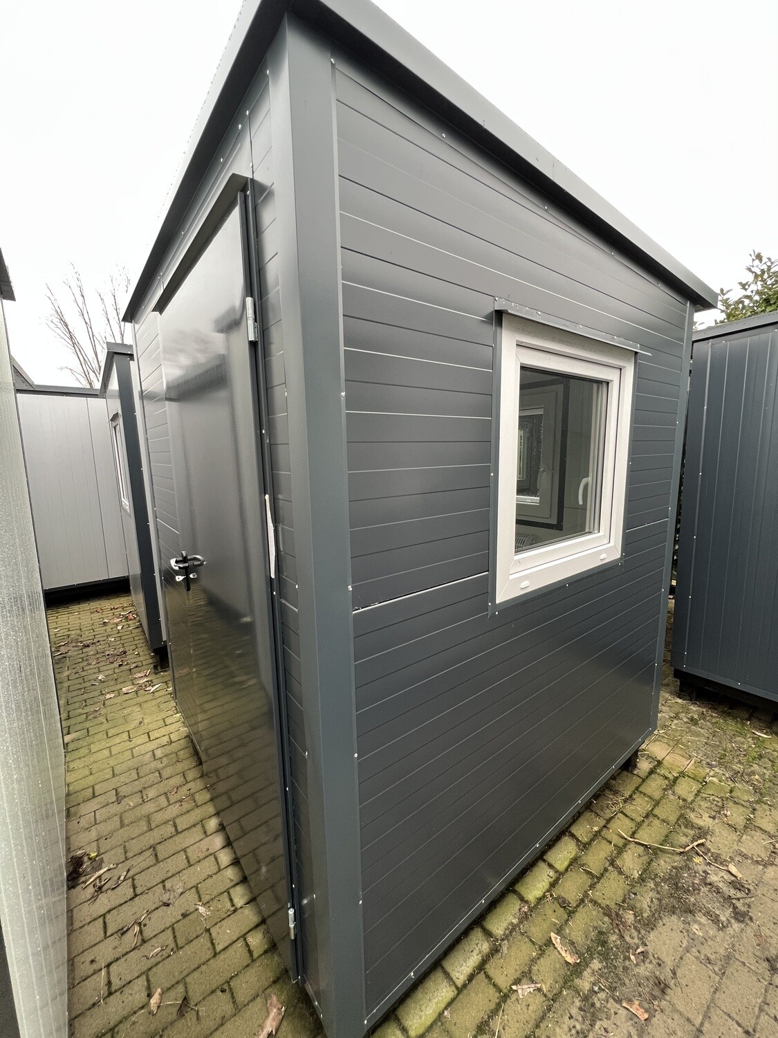 Dunkelgrau verkleideter Bürocontainer mit geöffneter Tür und Fenster, zwischen weiteren Containern