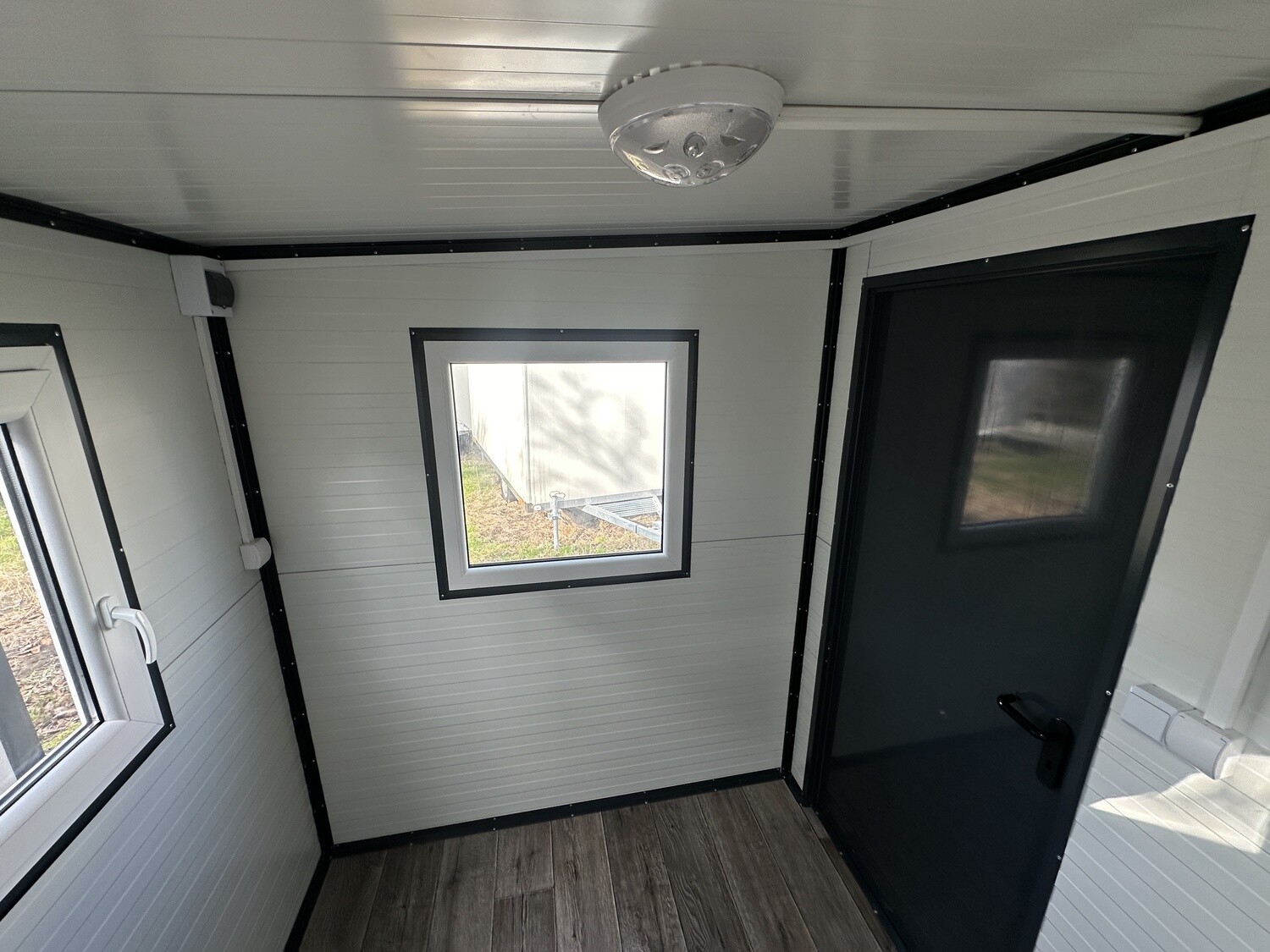 Innenraum mit Fenster, dunkler Eingangstür mit Sichtfeld und Deckenleuchte über Holzoptik-Boden