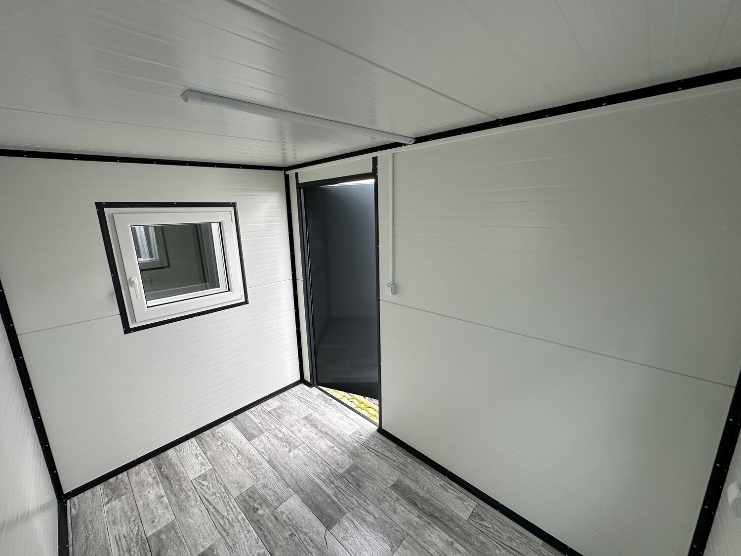 Blick durch den Innenraum zur offenen Tür, links daneben ein Fenster und graue Holzoptikdielen