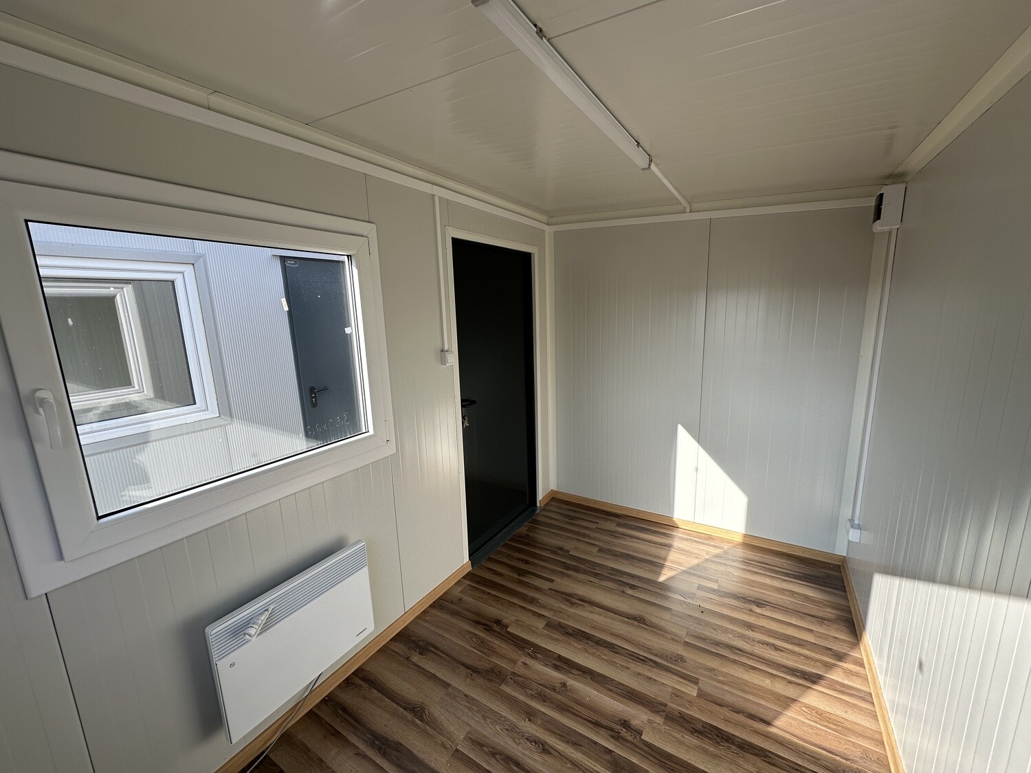 Breites Fenster mit Heizgerät darunter, daneben die dunkle Tür des Bürocontainers 3,50 x 2,20 m