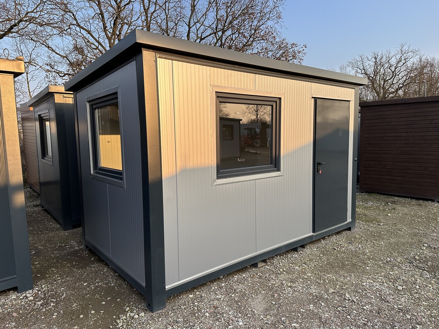 Hell verkleideter Bürocontainer 4,00 x 2,20 m mit dunklem Rahmen, Fenster und Tür auf Kiesfläche