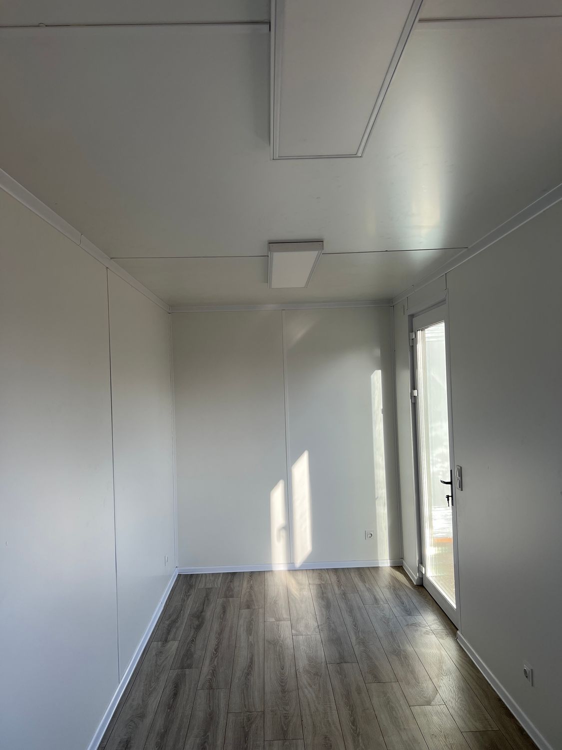 Blick zur verglasten Eingangstür im Bürocontainer 6,00 x 2,44 mit hellen Wänden und Holzdekorboden