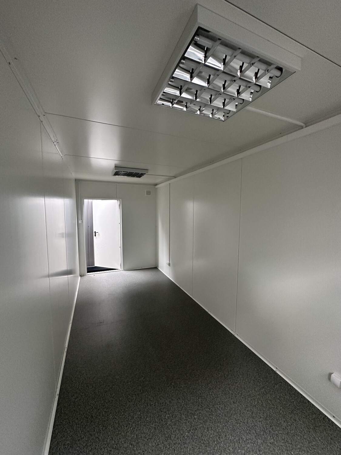 Leerer Büroraum mit Blick zur geöffneten Stirntür, Rasterleuchte und Lüftungsgitter an der Decke