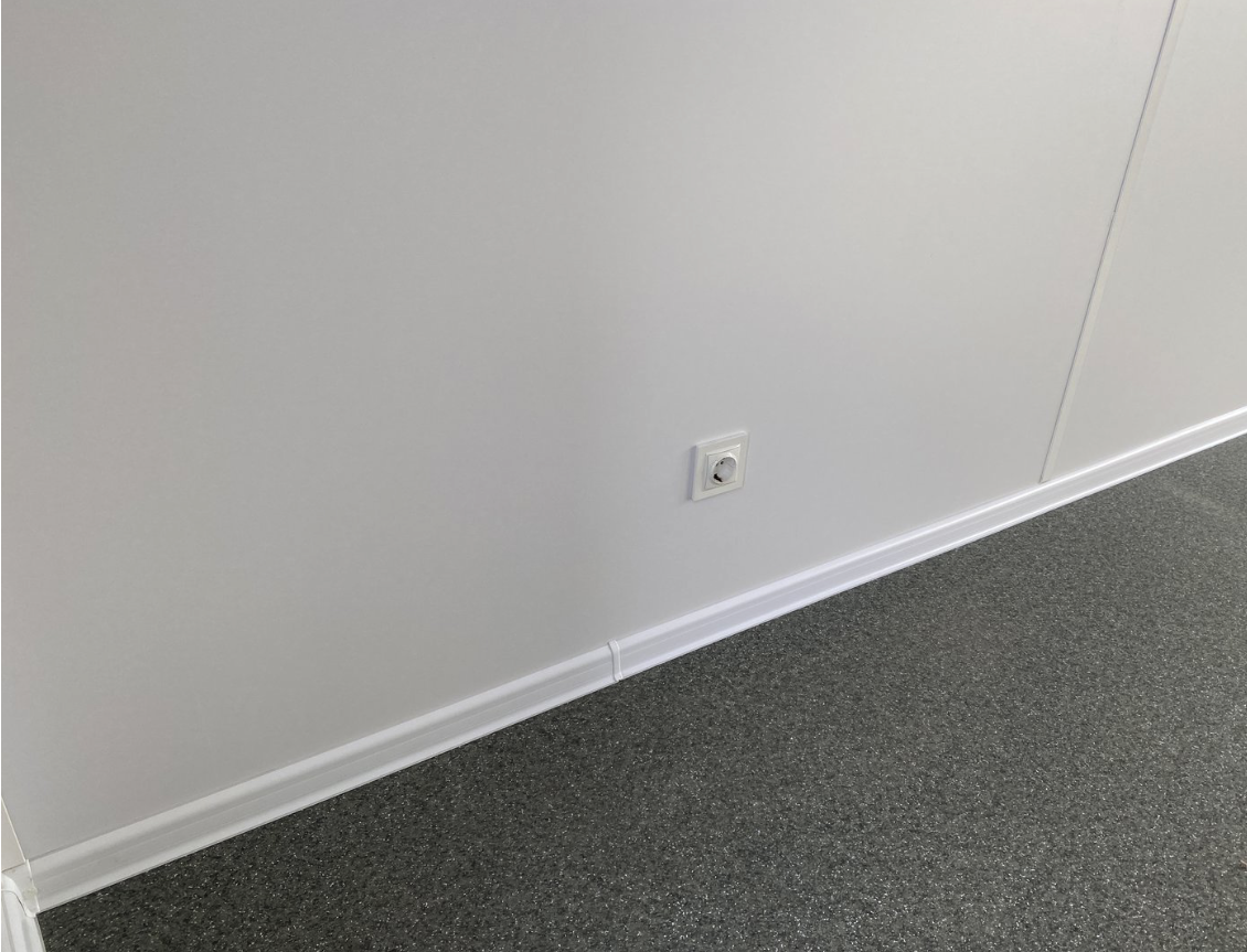 Detailaufnahme einer Steckdose an heller Wandverkleidung über grau gesprenkeltem Bodenbelag