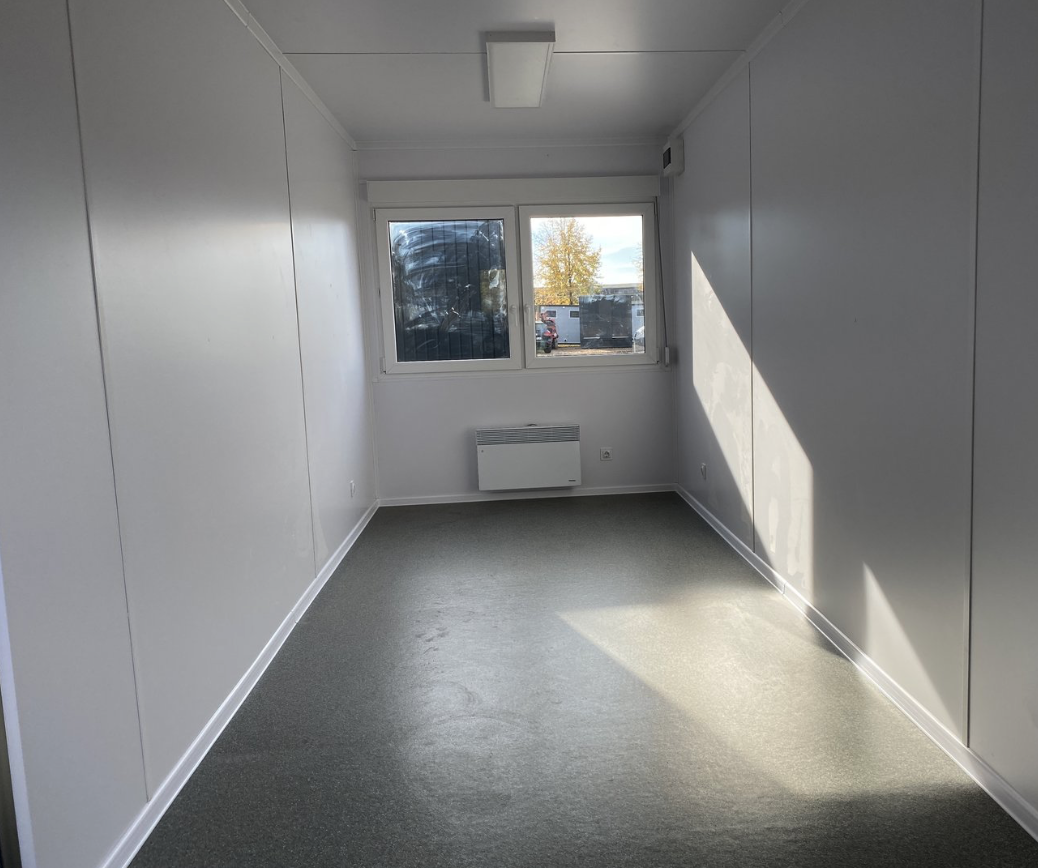 Innenraum des 6,00 x 2,44 m Bürocontainers mit grauem Bodenbelag, Fenster und Heizgerät
