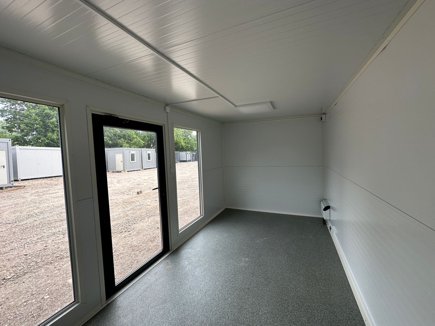 Grau melierter Bodenbelag, weiße Wandverkleidung und dunkel gerahmte Glastür im Bürocontainer