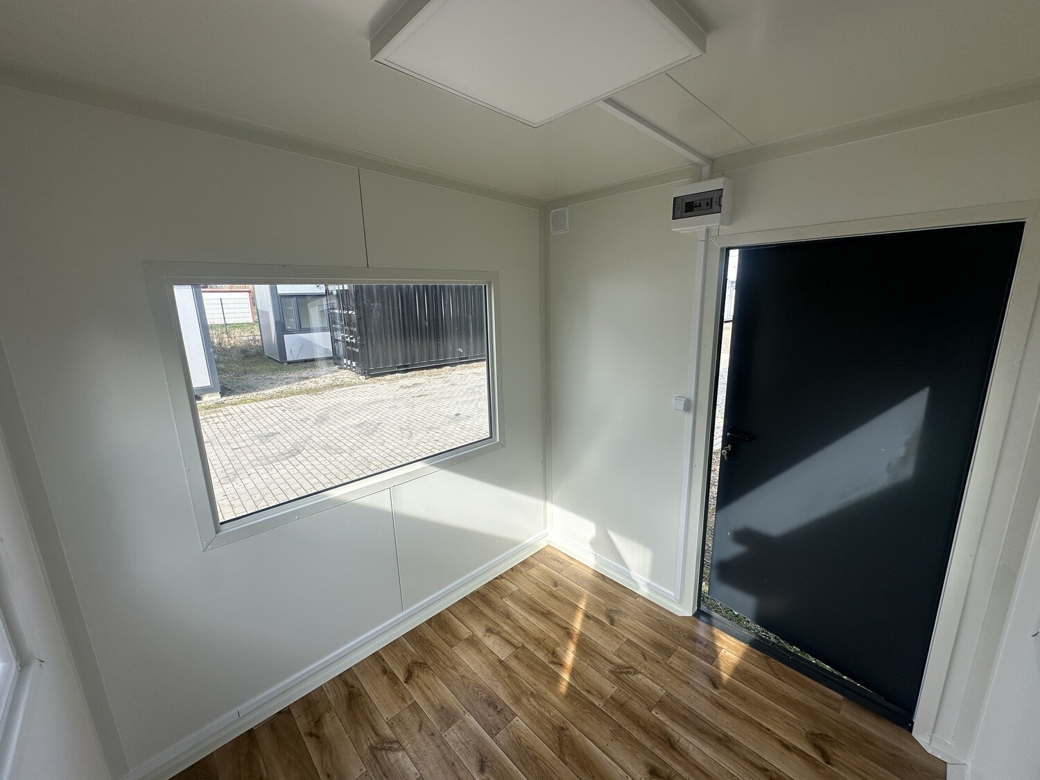 Blick zur geöffneten dunklen Eingangstür, daneben Fenster, Deckenleuchte und Boden in Holzoptik