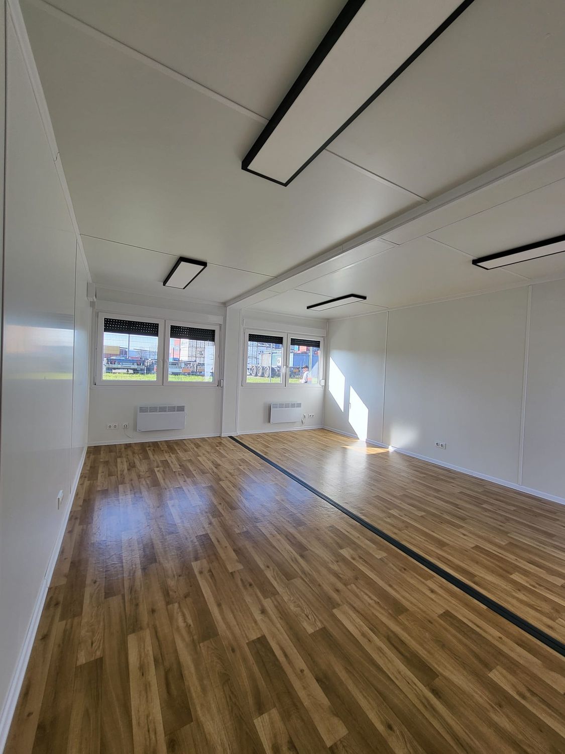 Innenraum mit holzfarbenem Bodenbelag, Fenstern mit Rollladen, Elektroheizkörpern und LED-Panelen