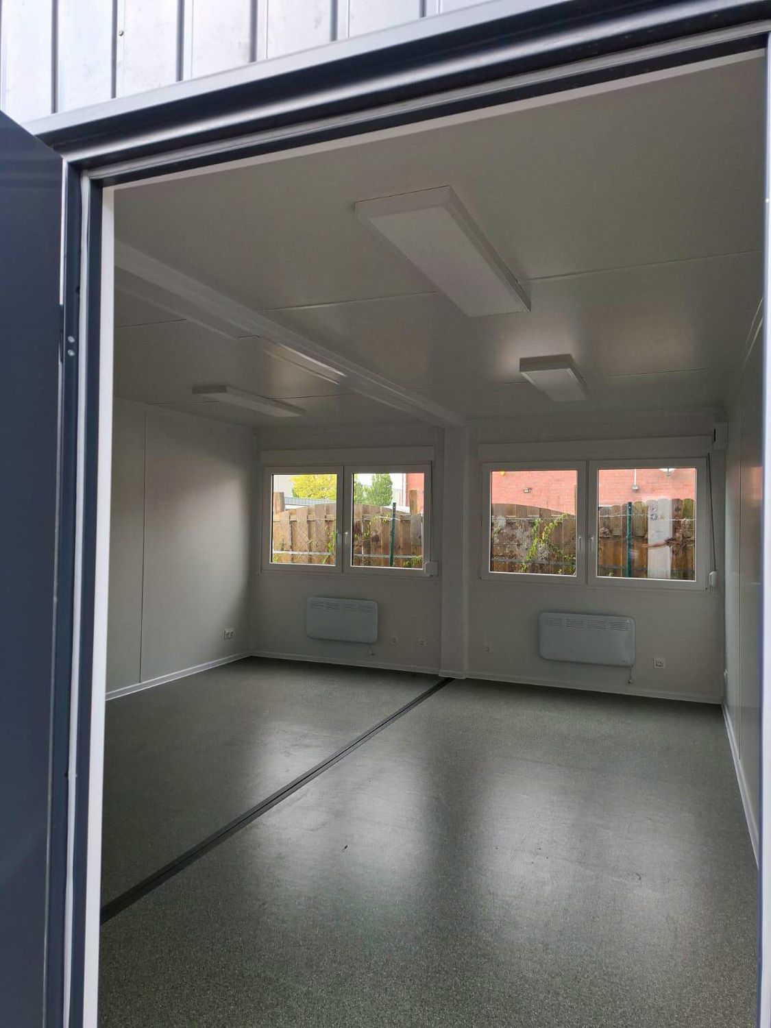 Durch die geöffnete Tür fällt der Blick auf grauen Boden, vier Fenster und zwei Wandkonvektoren