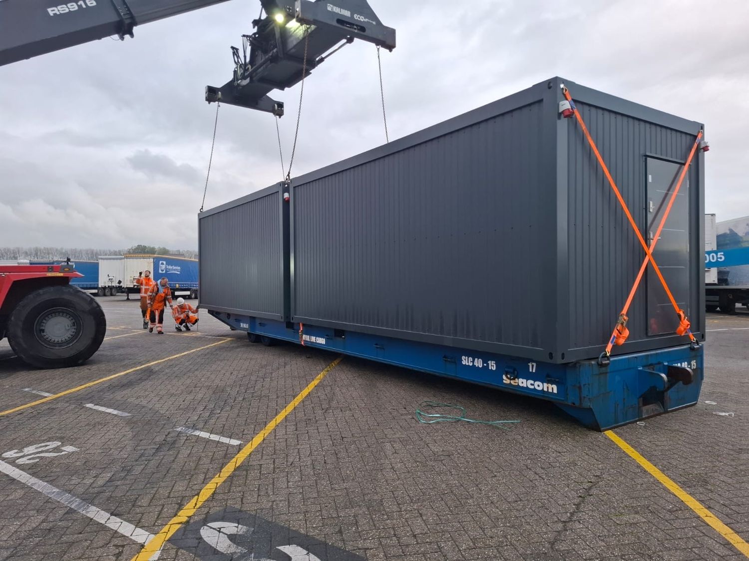 Verladung eines langen anthrazitfarbenen Containers per Kran, Mitarbeiter in Warnkleidung daneben
