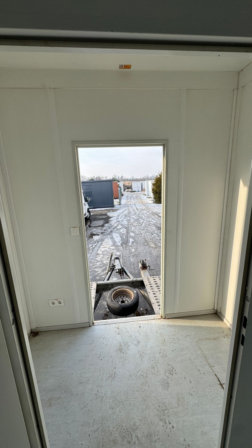 Aus dem Innenraum durch die geöffnete Außentür auf den Hof mit Anhänger und Containern