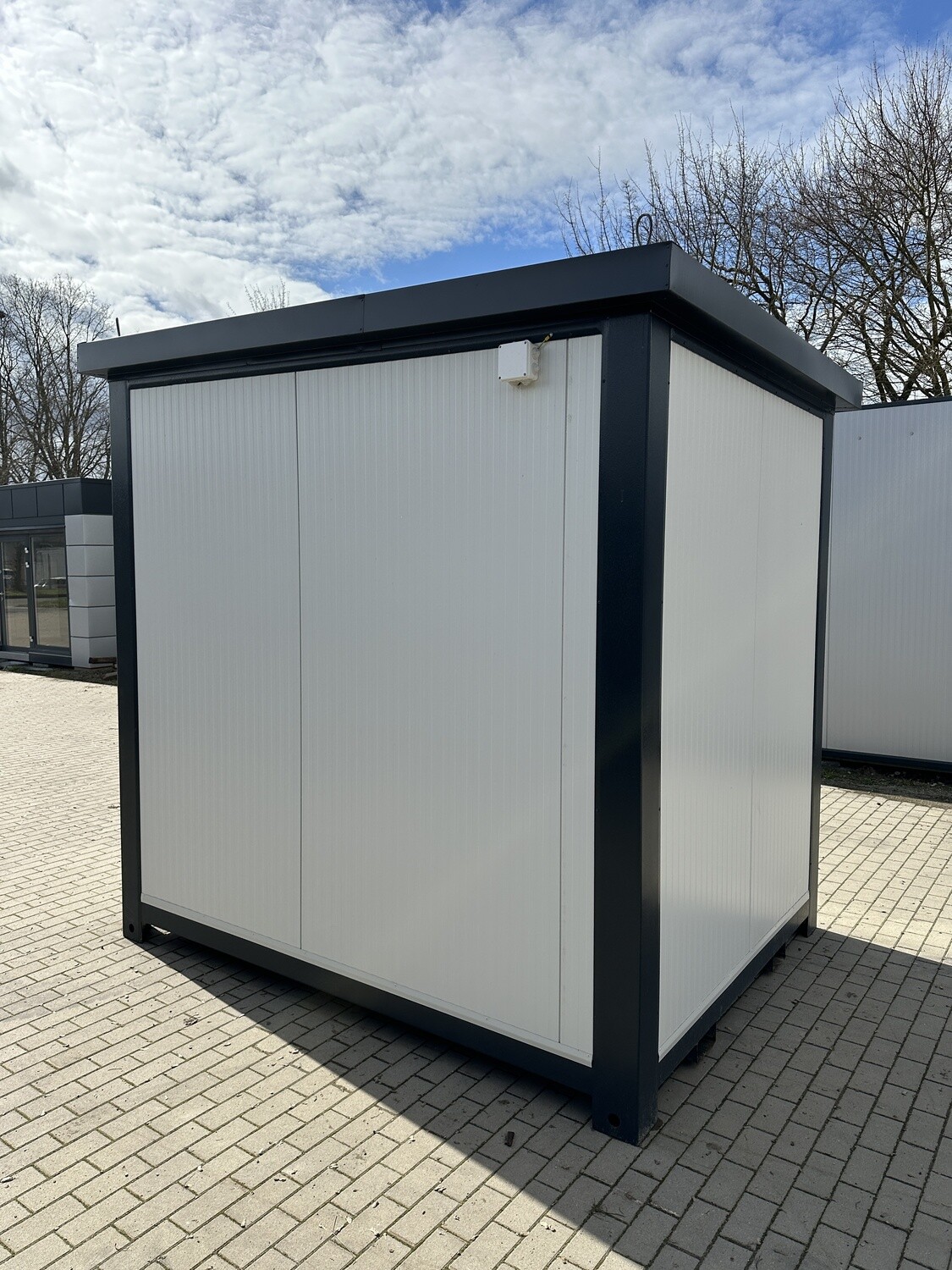 Seitenansicht des Lagercontainers auf gepflastertem Platz, weiße Wand mit Elektrokasten am Dachrand