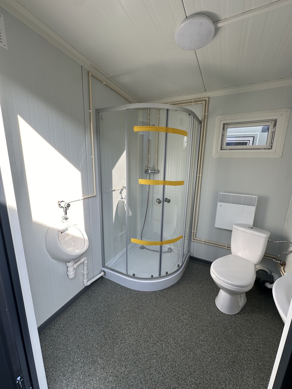 Runddusche, Urinal und WC im Innenraum, dazu Wandheizung und grau gesprenkelter Bodenbelag