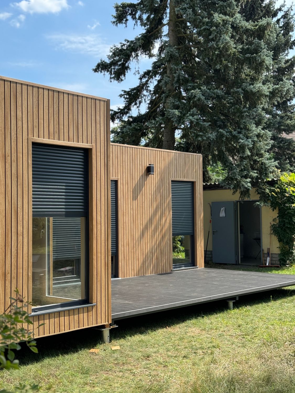 Seitlicher Blick entlang der Holzfassade des Wochenendhauses: bodentiefes Fenster, Rollladen, Holzterrasse
