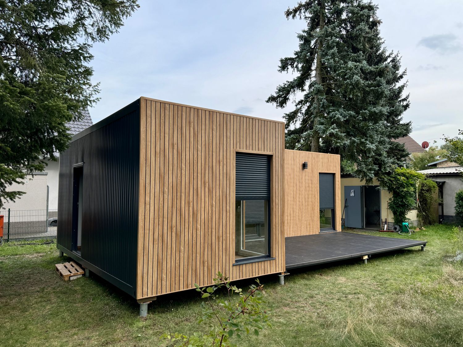 Von der schwarzen Stirnseite aus zeigt sich das Tiny House in L-Form mit Holzverkleidung und Terrassendeck