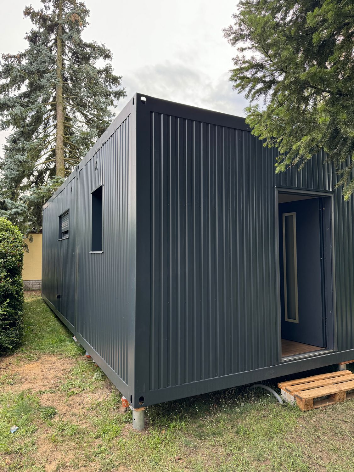 Noch ohne Holzverkleidung: anthrazitfarbene Containerseite mit offener Tür, Fenster und Stapelecken