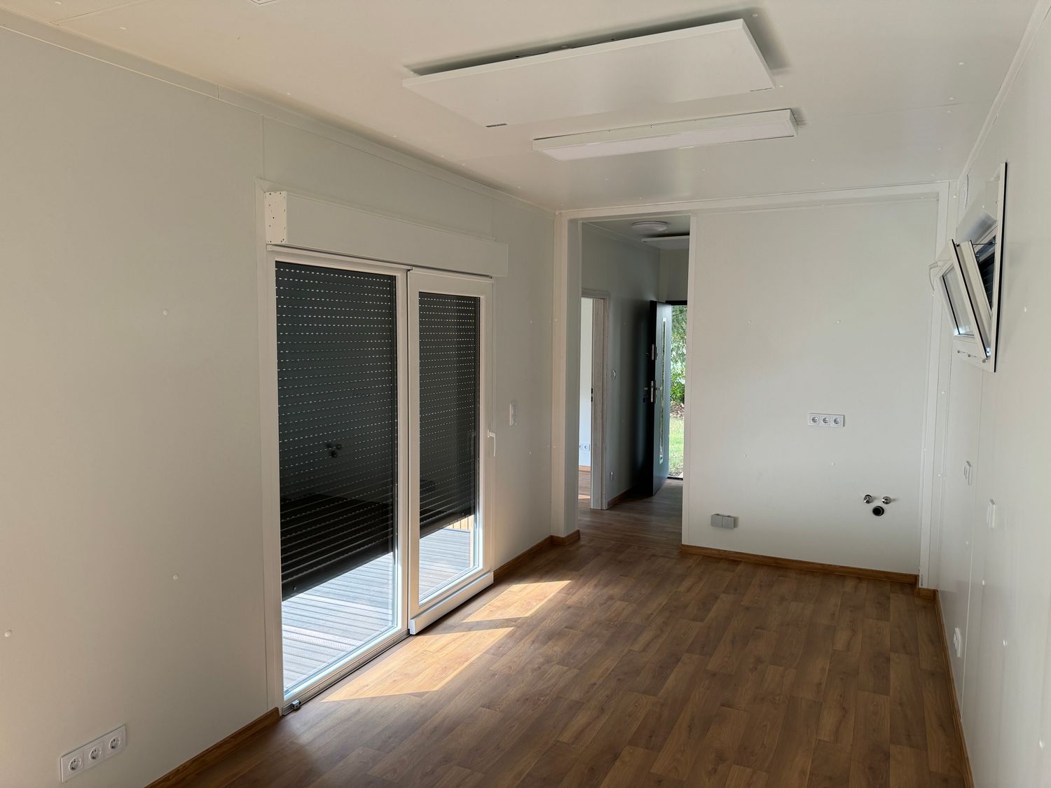 Wohnraum mit Blick in den Flur: Terrassen-Schiebetür, Deckenpaneele und Steckdosen an der hellen Wand