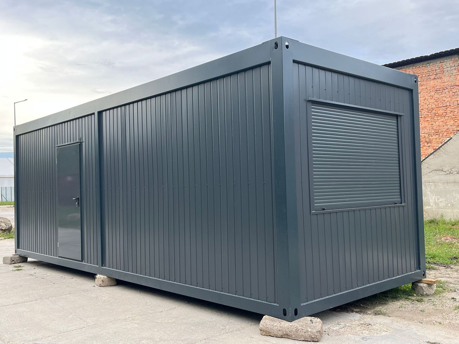 Eckansicht des Wohncontainers mit heruntergelassenem Rollladen am Stirnfenster und Seitentür