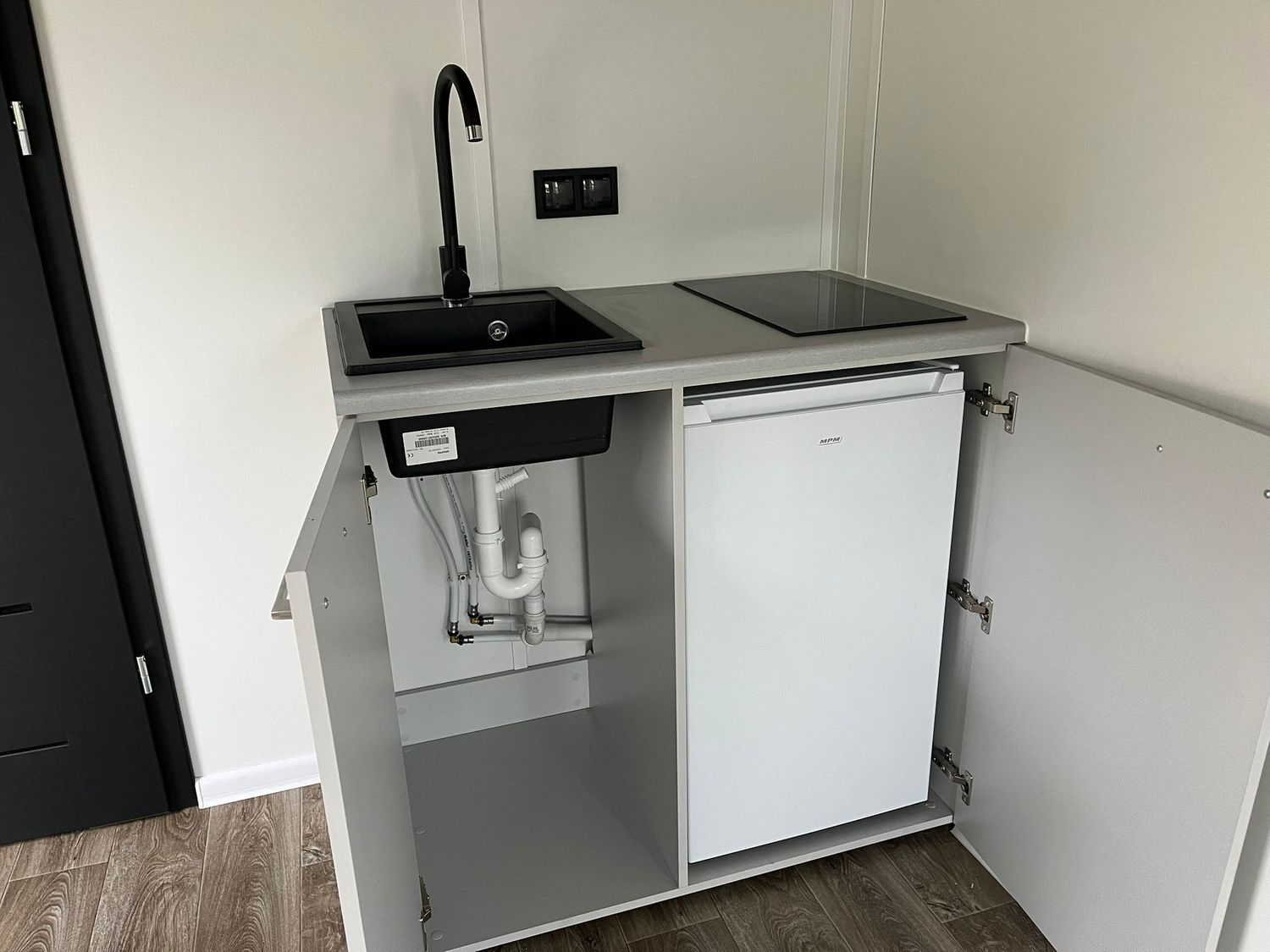 Detailaufnahme der geöffneten Unterschränke mit Siphon, Kühlschrank, Spüle und Kochfeld