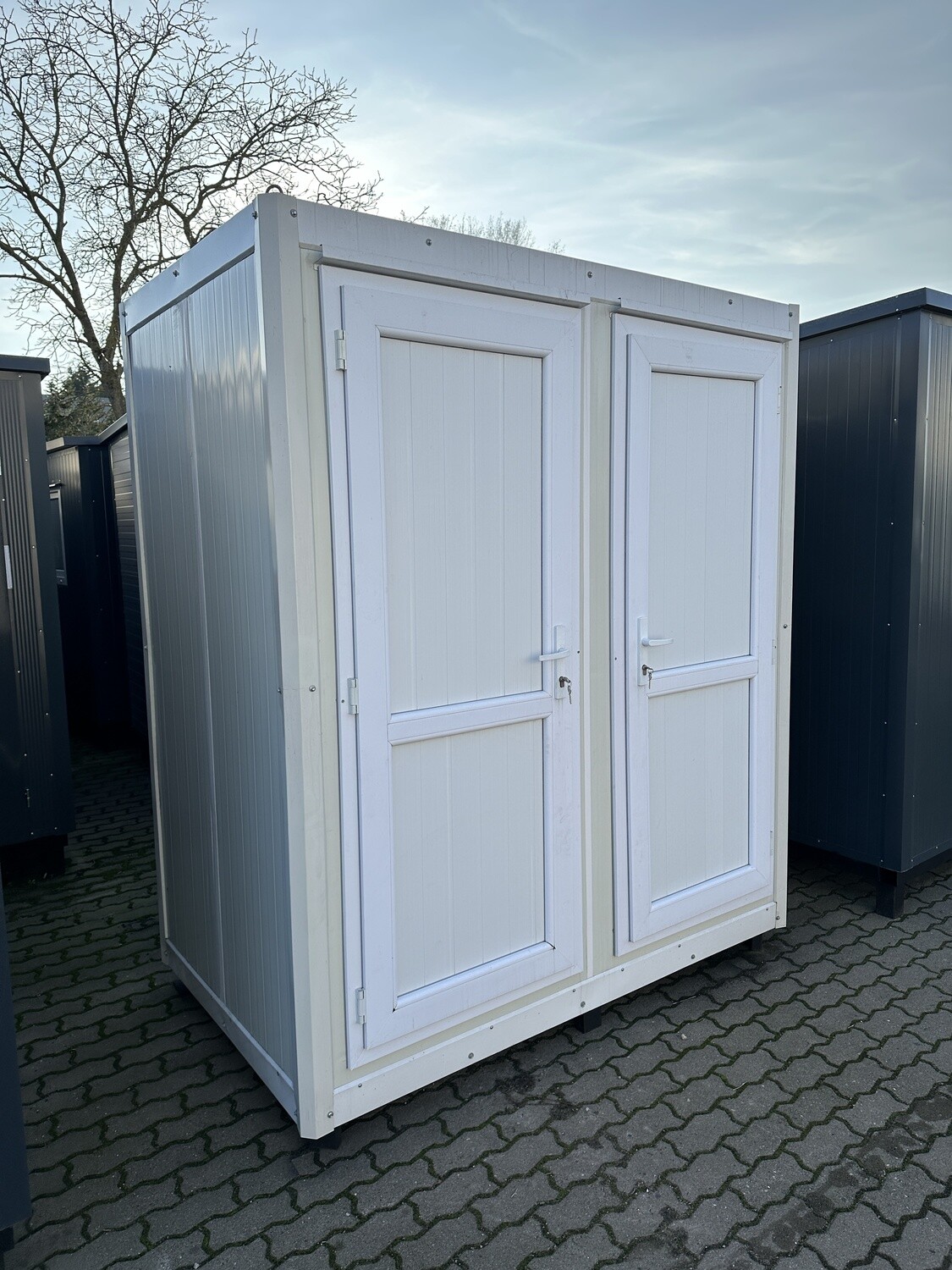 Schrägansicht des Sanitärcontainers mit zwei geschlossenen weißen Türen auf Pflasterfläche