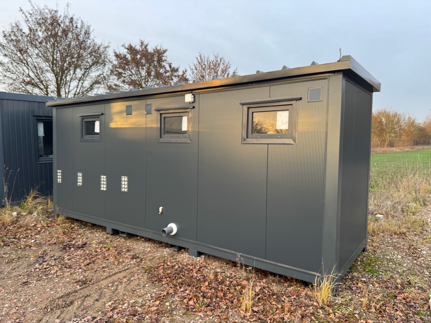 Rückseite des Sanitärcontainers 6,00 x 2,00 m mit drei Fenstern, Lüftungsgitter und Abflussrohr