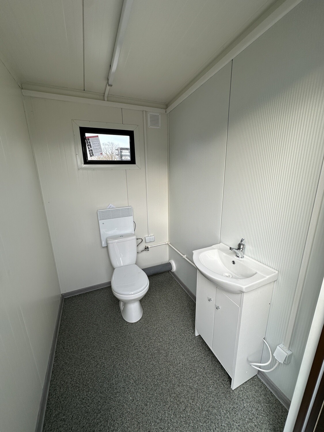 Einzelabteil von innen mit WC, Wandheizgerät, Waschtisch und kleinem Fenster, grauer Boden
